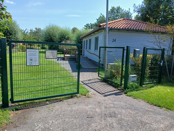 Im Vordergrund ist ein grünes Tor zu sehen, dessen rechter Flügel offen steht, im Hintergrund ist links ein weiß gestrichenes Holzhaus zu sehen
